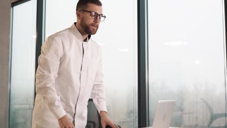 Médico-Masculino-Enfocado-Se-Sienta-En-Un-Escritorio-En-Una-Clínica-Moderna-Con-Ventanas-Panorámicas,-Mira-Una-Computadora-Portátil-Y-Reflexiona