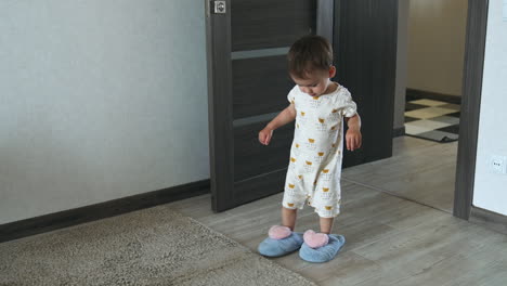adorable baby stands near the fluffy slippers. kid puts on home footwear and runs in them by the carpet.