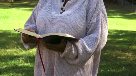 medium shot of black woman standing in grass on a sunny day reading her bible and spending time with god no face