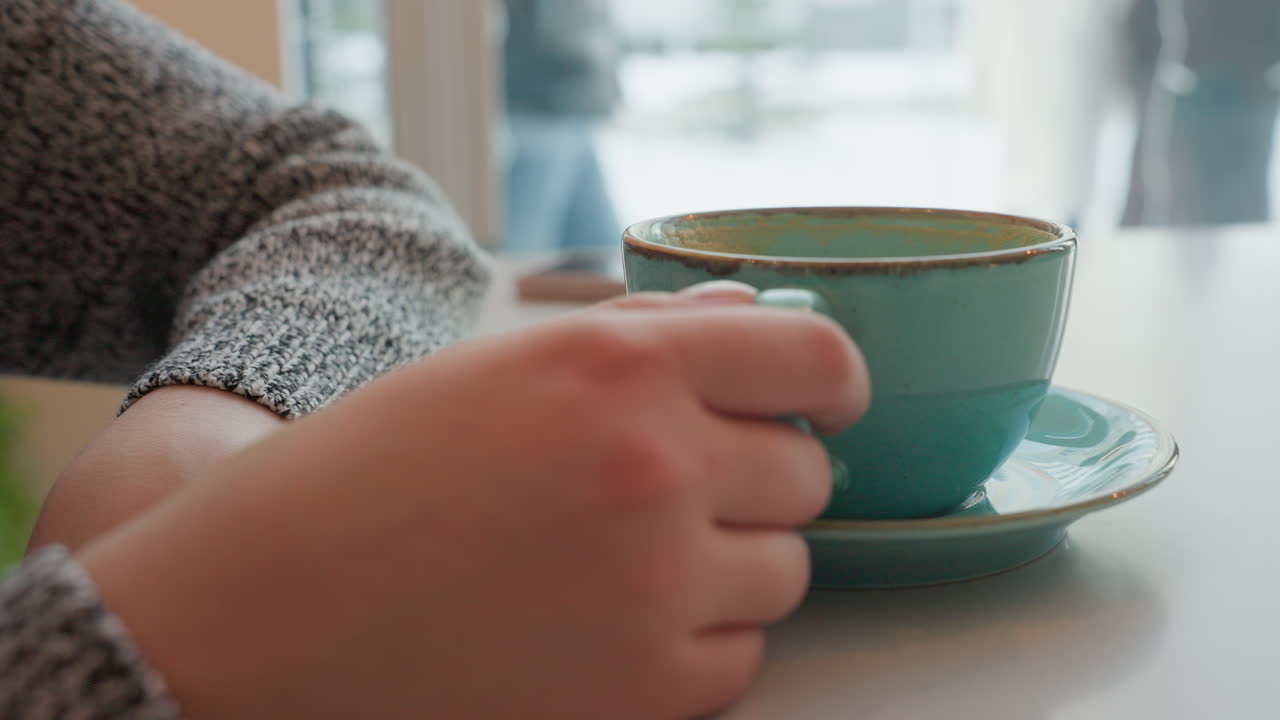 Freelancer holding cup of hot coffee while sitting inside restaurant during winter as people outside discuss in snowy environment visible through window, creating warm peaceful indoor atmosphere