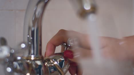 female hand turning knob on tap and pouring water into glass in bath room