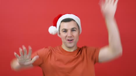 a young man in an orange t-shirt and santa claus hat dances waving his arms. the guy in red christmas outfit is standing in front of a red background