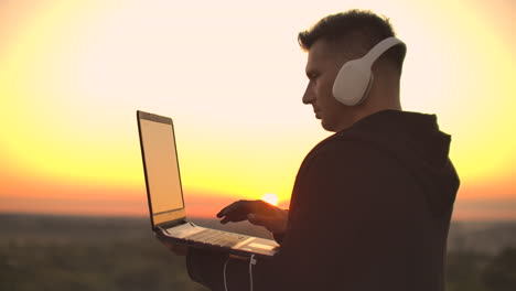 a man in headphones on the roof relaxes working remotely enjoying life despite a handsome kind of sipping beer and types on the keyboard. trade on the stock exchange using a laptop and enjoying the beautiful view