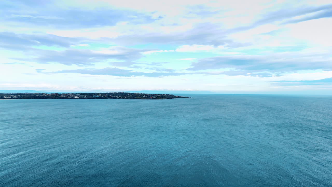 Vista diurna do oceano e da costa. A água se estende pela cena enquanto as ondas rolam perto da costa. O céu tem nuvens que espalham luz sobre o oceano