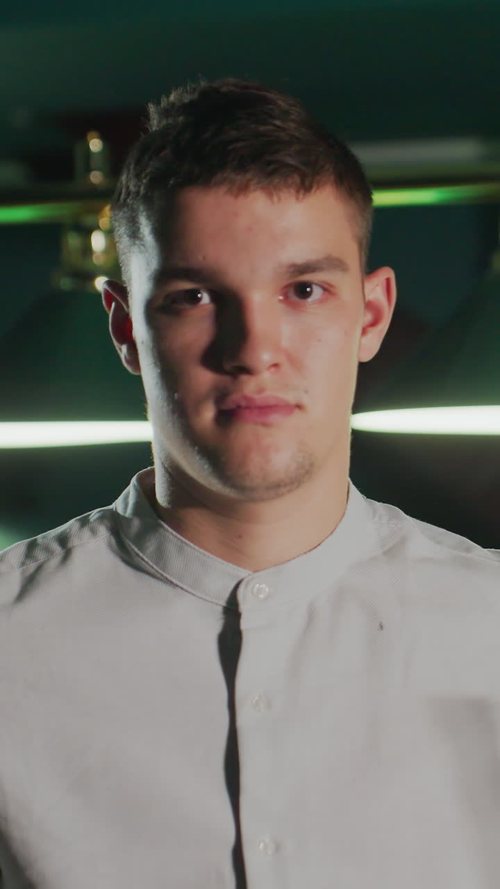 셔츠를 입은 남자가 진지한 표정으로 희미하게 조명된 수영장 홀에 서기 전에 큐 스틱을 들고 앉아 있습니다. 집중된 시선과 강렬한 분위기는 경쟁 정신과 집중력을 강조합니다.