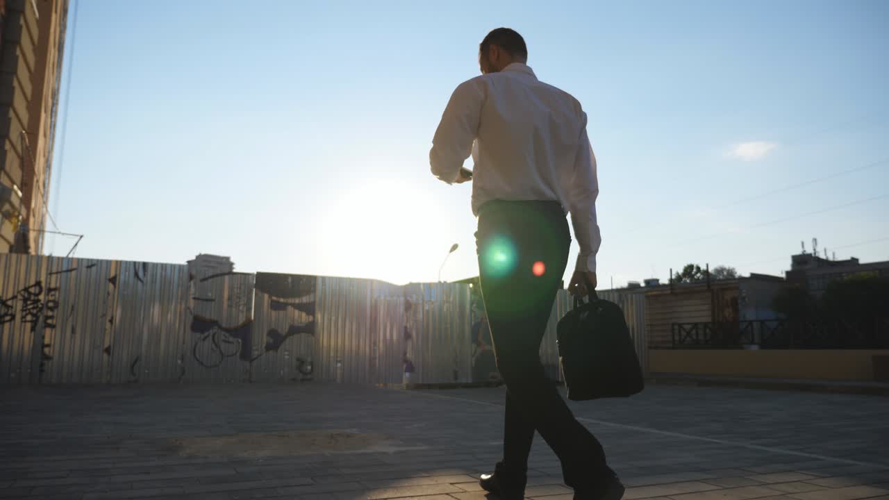 un hombre de negocios exitoso con ropa formal usa un teléfono inteligente yendo a trabajar en la acera. un joven empresario con un maletín navega por el teléfono móvil caminando cerca de un edificio de oficinas en una calle urbana. vista trasera en cámara lenta
