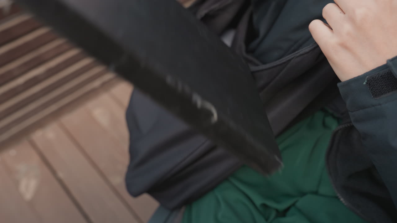 Una mano che fruga in una borsa a tracolla nera, estrae un quaderno e un tablet mentre è seduta su una panchina di legno in un ambiente urbano all'aperto, preparazione concentrata per una riunione o uno studio, cerniera e tracolla ben visibili