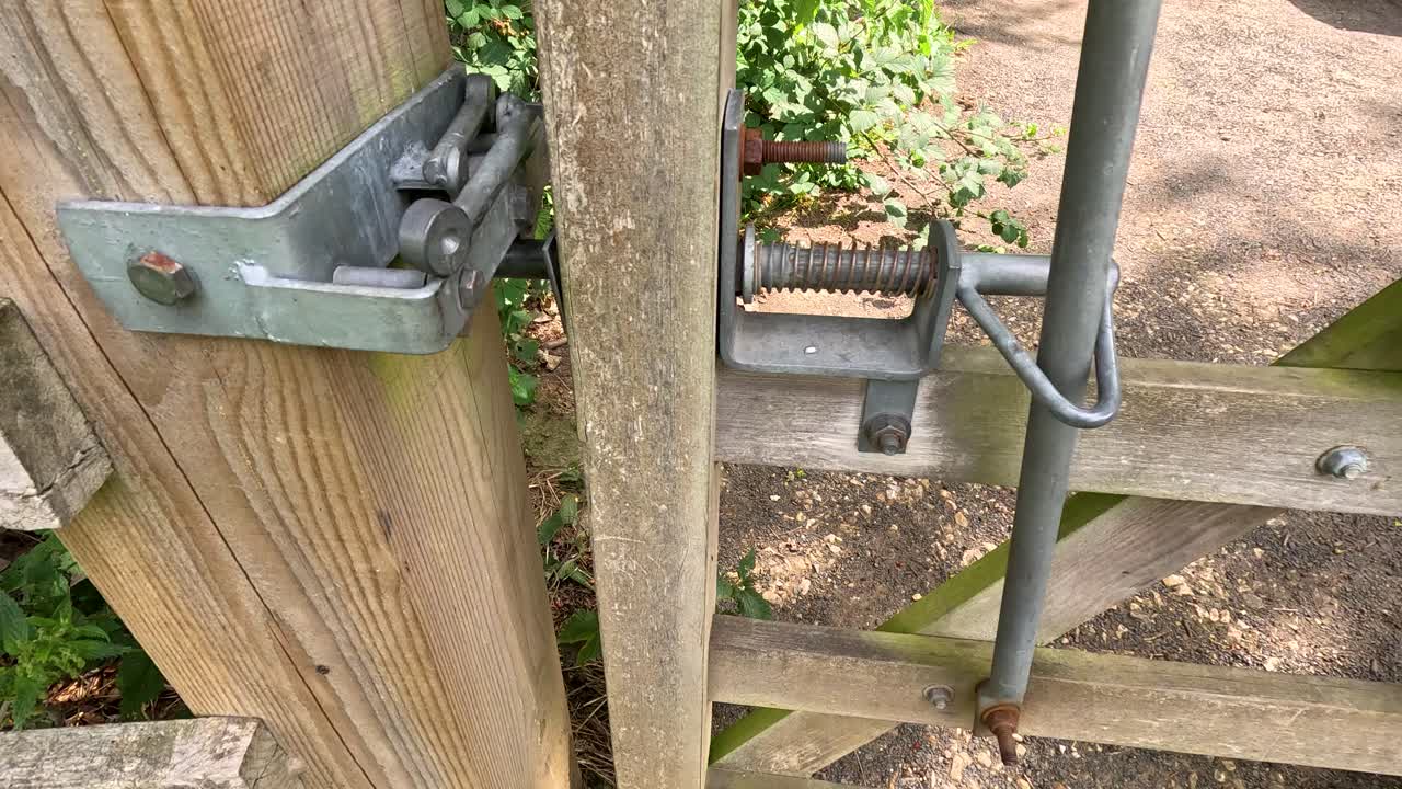 A close-up sequence shows a metal latch mechanism securing a wooden gate, with natural daylight and garden foliage visible in the background. Camera remains steady