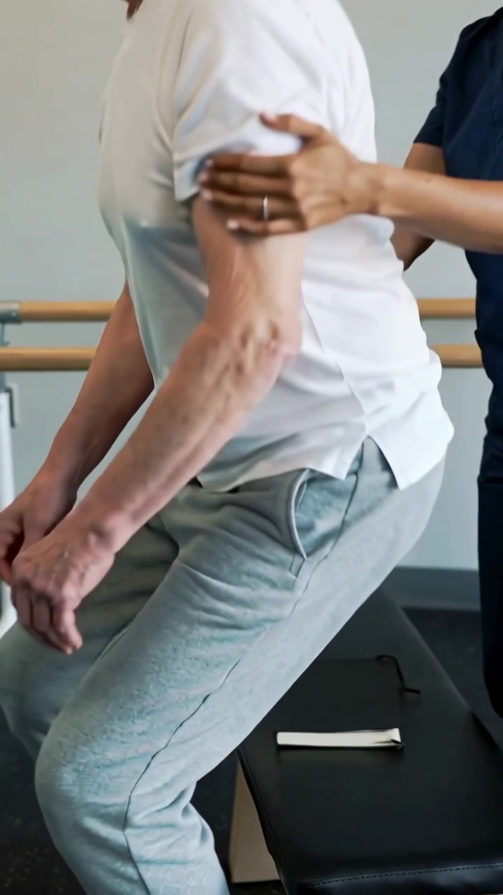 Vertikal video: Plasserer hånden, kliniker veileder eldre mann til å stå på klinikk, hvit t-skjorte, briller i nærheten.
