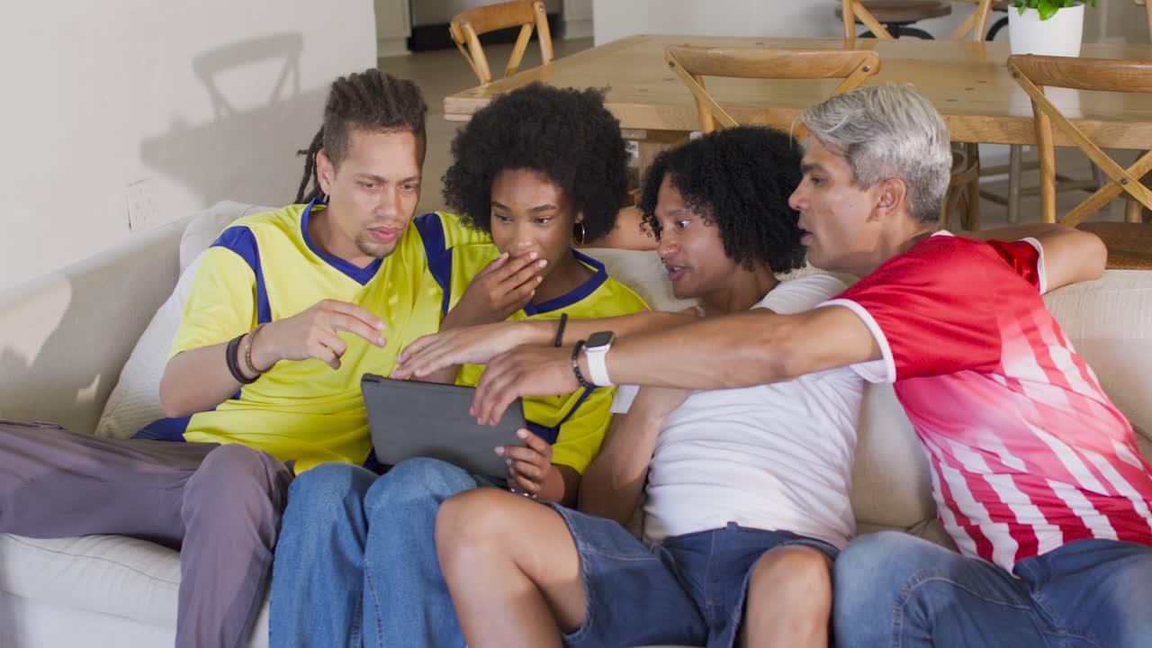 African American friends in jerseys on sofa leaning toward tablet pointing tapping reacting to clip