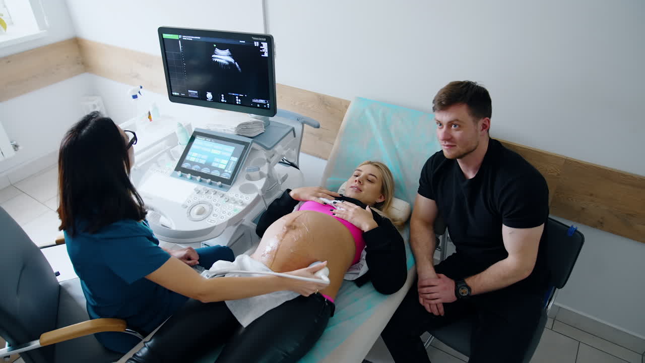 Expecting parents are in the doctor's cabinet. Woman is undergoing ultrasound check up. Top view.