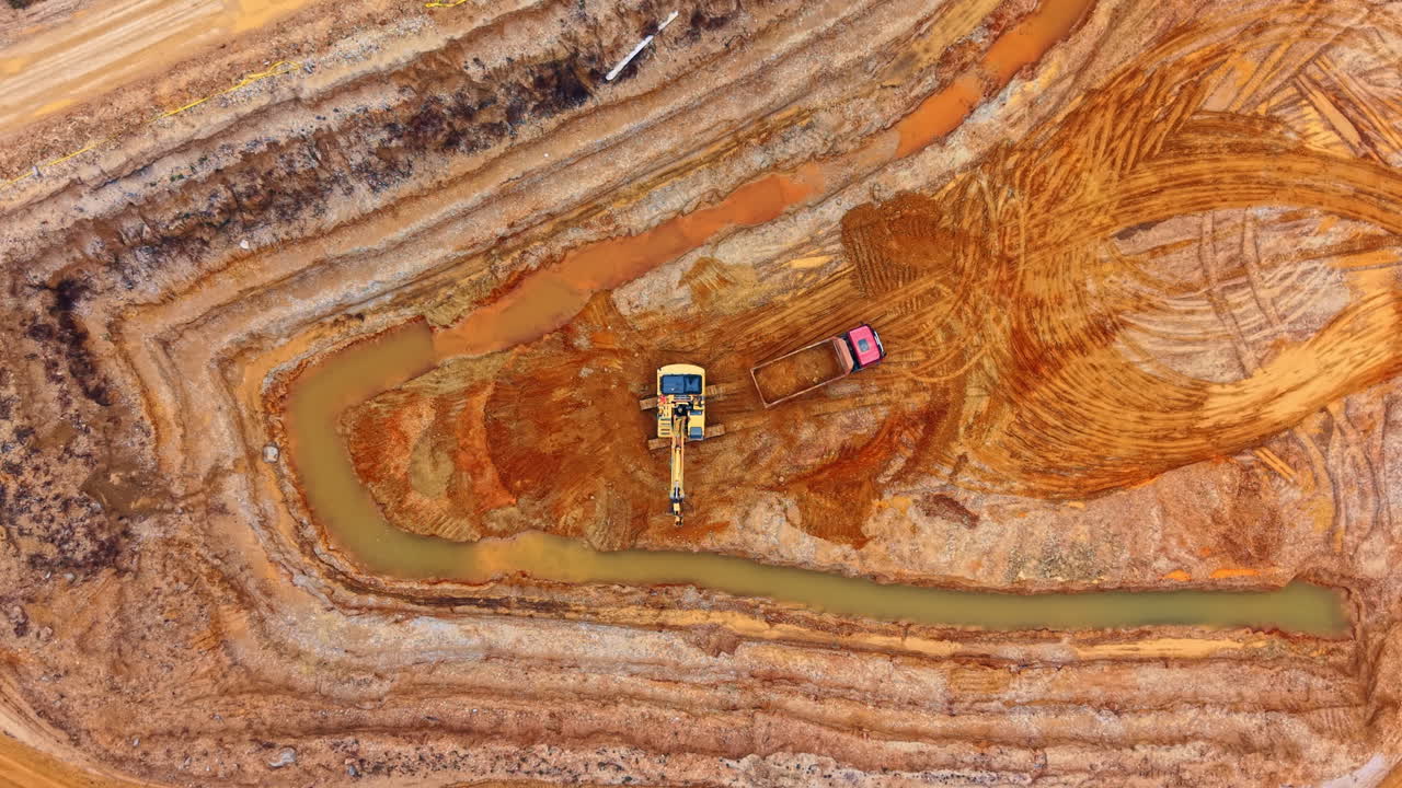 Excavator loading sand into dump truck. Aerial top down view of yellow excavator and red truck working in sandy quarry pit with water channels