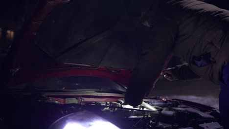 man looking at engine of broken down car with flashlight at night. headlights turned on