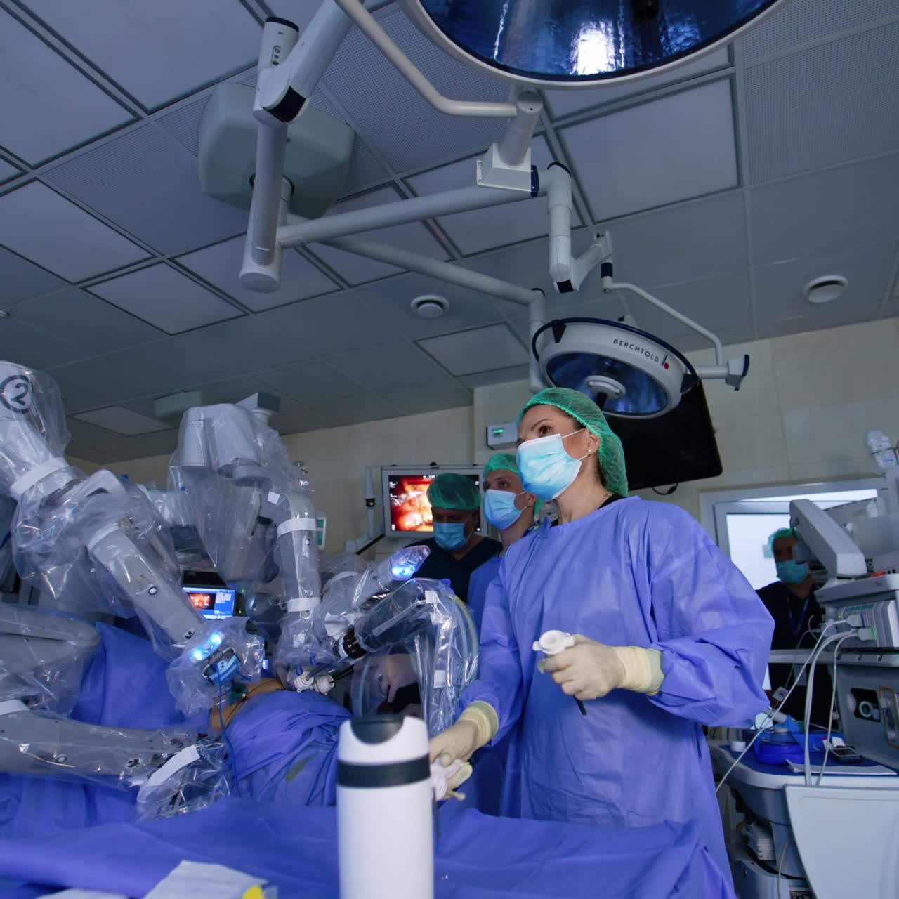 Doctors' team work in a surgery room with robotic surgeon. Arms of Da Vinci machine move performing laparoscopy. People look at monitors intently. Low angle view