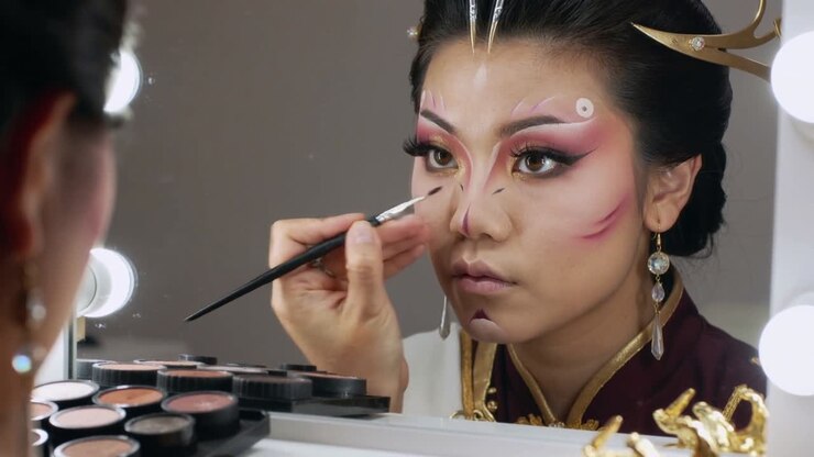 Asiatisk performer lægger scenemakeup ved oplyst spejl, holder børste klar, med palet og bordeaux krave.