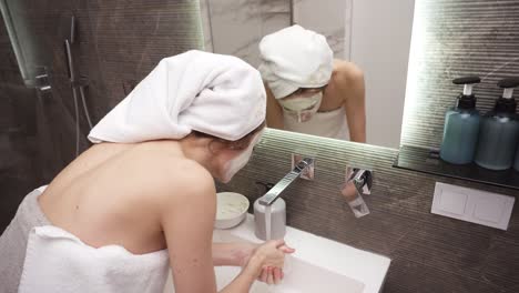 a woman standing in the bathroom near the sink in white towel washes off a white cosmetic mask with water from the tap. rare view