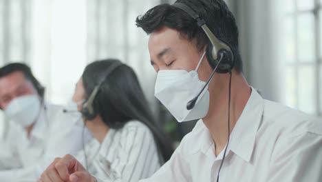 close up of a man of three asian call center agents wearing headset and mask headache because two of his colleagues are talking during working at the office