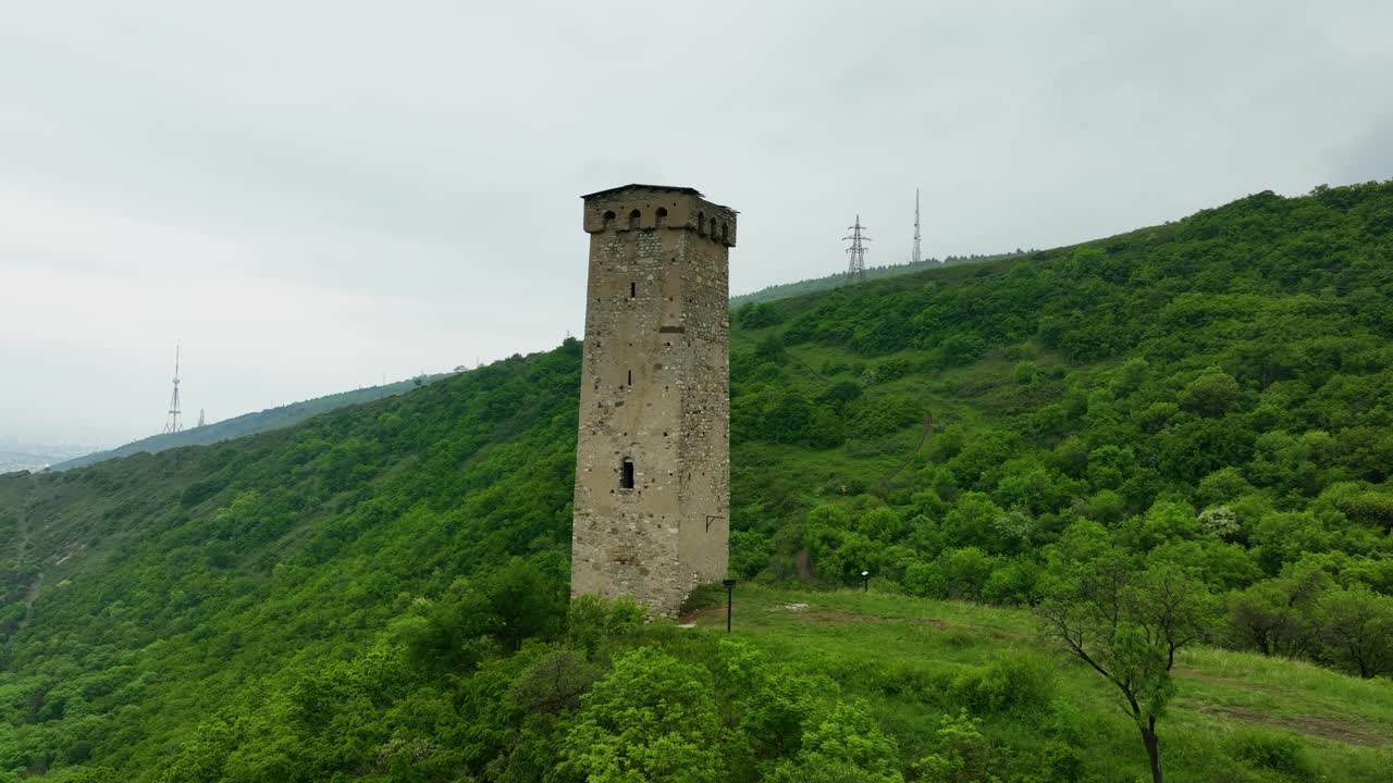 Drone view of a historic watchtower standing alone among the vivid green hills of Tbilisi’s outskirts under soft daylight