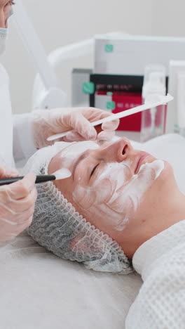 beautician applying peeling moisturizing cream mask on young woman face for rehydrate face skin