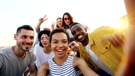punto de vista de una chica afroamericana sosteniendo una cámara y tomando selfies con amigos felices en una fiesta en el techo. hombres y mujeres están mirando a la cámara, posando y riendo.