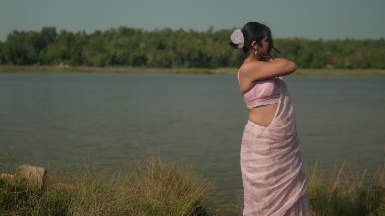 Wanita bersaree merah muda menikmati semilir angin di tepi sungai, dengan rumput di latar depan, pada hari yang cerah.