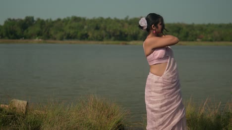 woman in pink saree enjoying breeze by river, grass in foreground, sunny day