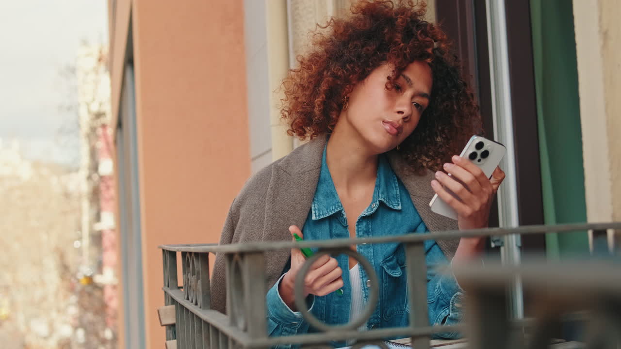 Giovane donna con capelli ricci usa lo smartphone sul balcone