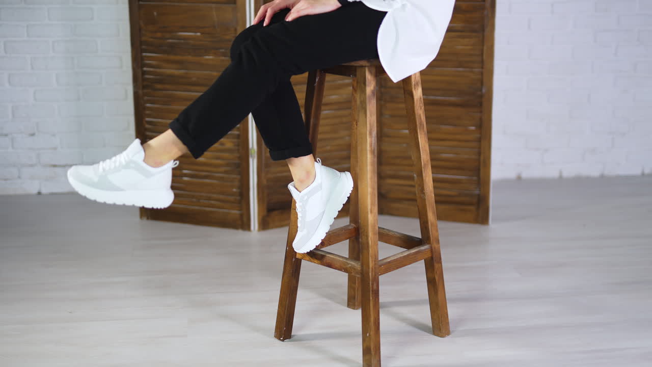 Woman sits on a high stool waving her feet in white sneakers. Model jumps off the tabouret and walks away in comfortable footwear.