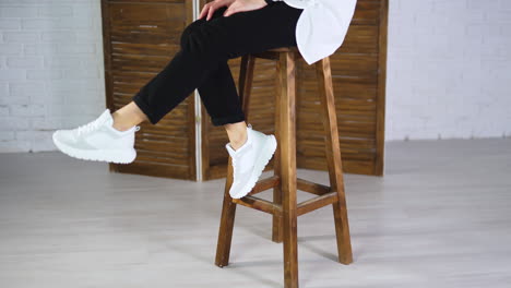 woman sits on a high stool waving her feet in white sneakers. model jumps off the tabouret and walks away in comfortable footwear.