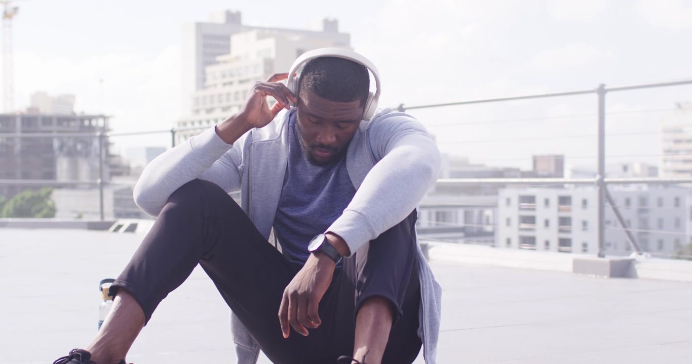 African American man adjusting headphones checking smartwatch on rooftop with skateboard behind