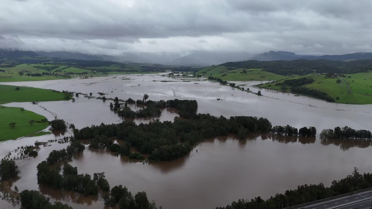 Gerringong under floodwaters with visible farmland, trees, and infrastructure affected by storm, panoramic establishing aerial drone