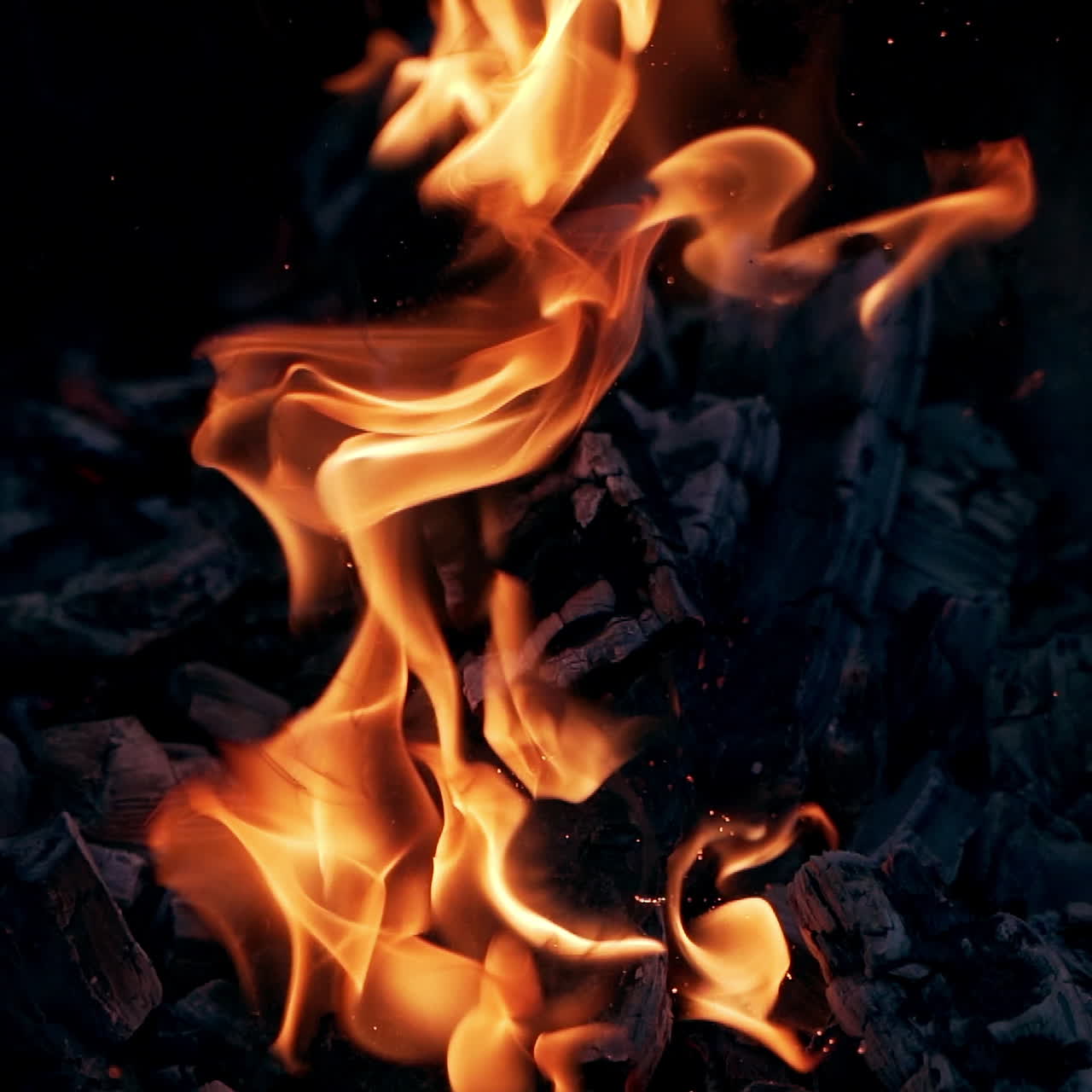 Explosion of flame with bright sparks. Black smoldered logs explode with fiery flame on dark background outdoors. Close-up.
