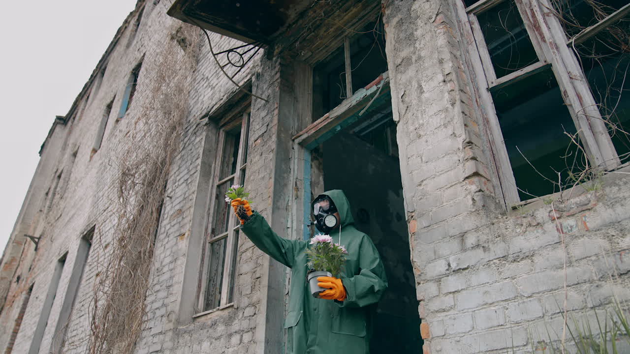 Ecologist standing with plants in abandoned place. Person in protective uniform and gas mask holding flowers near the destroyed building and looking on it. Chemical attack and air pollution concept