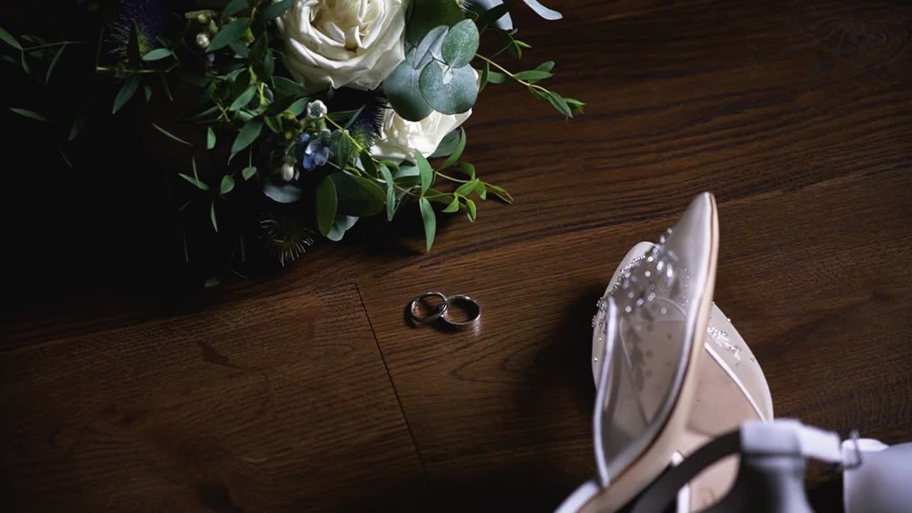 Top view of the wedding rings, bridal shoes and flower bouquet on a dark wooden surface, rotating shot