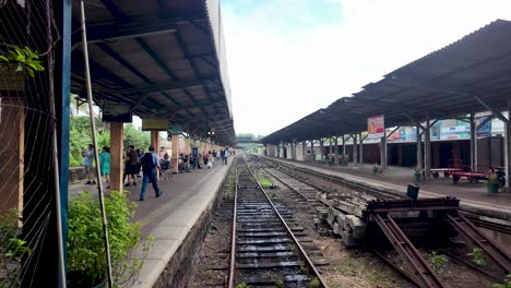 kandy railway station in sri lanka, where people are gathered on the platform waiting for their train. the empty tracks stretch into the distance under a covered roof.