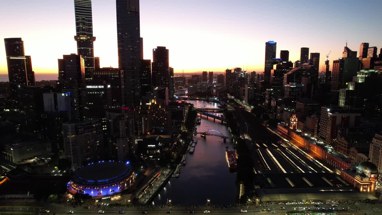 Volo aereo lungo il fiume Yarra a Melbourne con le luci della città al tramonto dorato.
