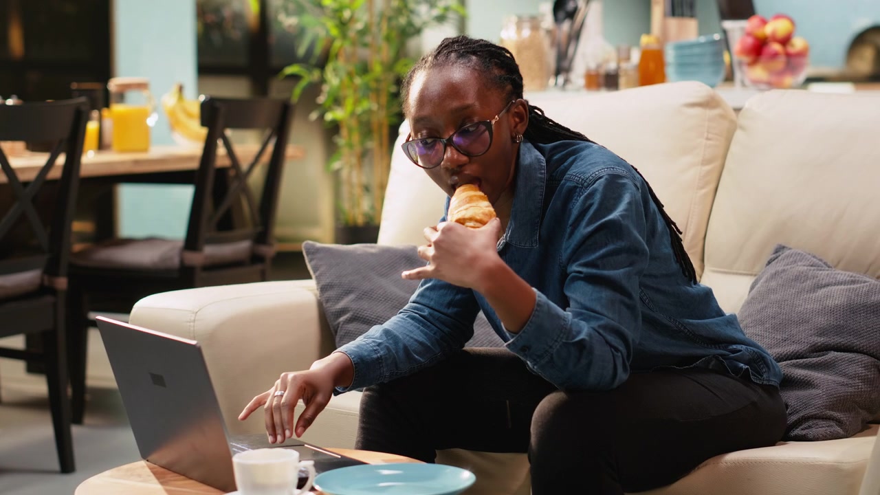 Young woman eating croissants and using laptop in home interior
