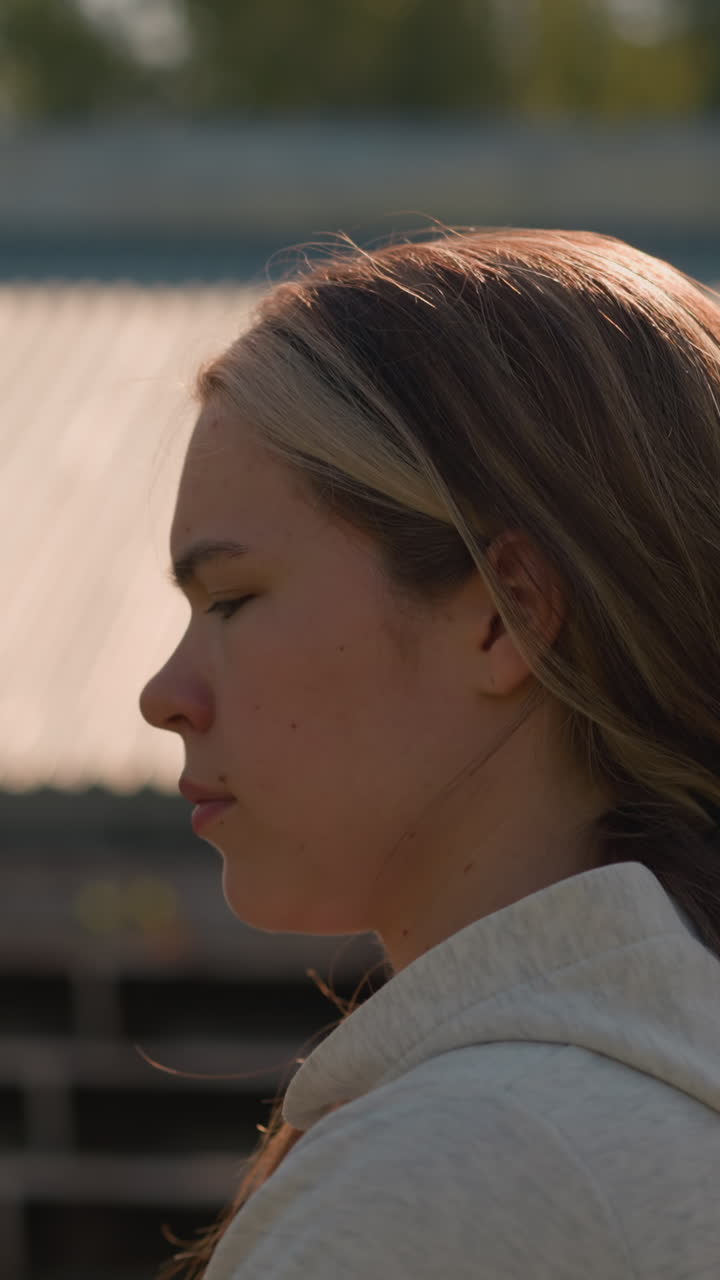 joven adulta con el cabello largo atado hacia atrás, con capucha, camina pensativa por las gradas vacías del estadio en un día soleado, parece reflexiva y profundamente pensativa