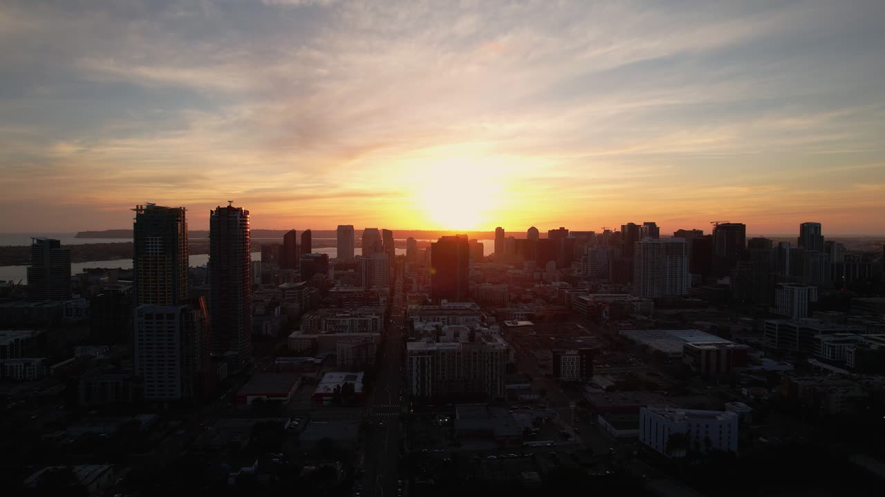 Drone voando para frente em direção ao horizonte do centro de San Diego ao pôr do sol com luz solar brilhante atrás dos edifícios.