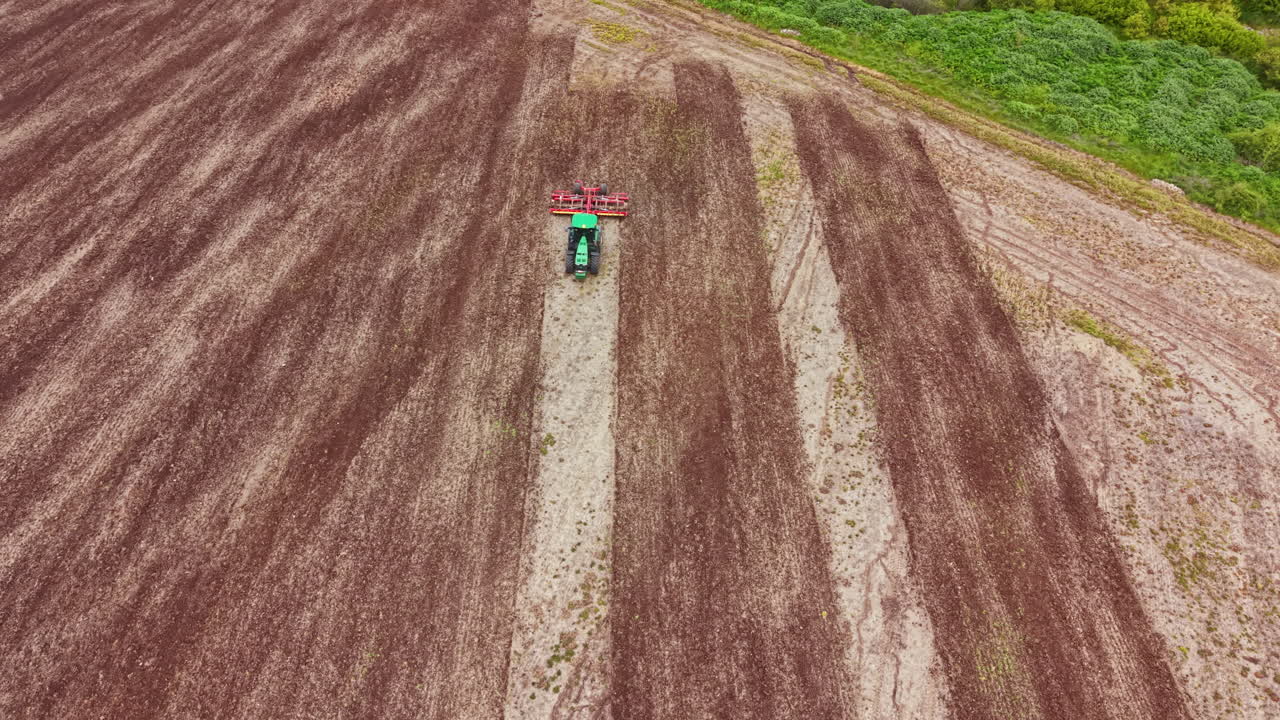 Un trattore lavora in un vasto campo per preparare il terreno alla semina, vicino a un'area boschiva, in pieno giorno.