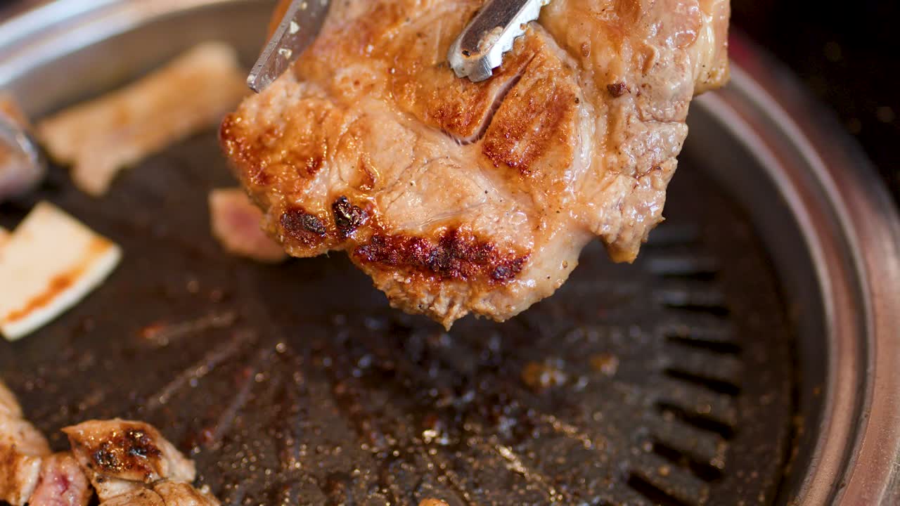 Hand flips pork steak with tongs on hot Korean barbecue grill, overhead view