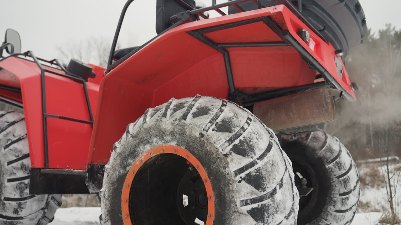 Pneumatici posteriori dell'ATV rosso che sollevano neve, primo piano del battistrada sovradimensionato e del cerchione arancione, vapore dallo scarico del motore, sospensioni robuste che si flettono, pilota che prepara una manovra fuoristrada in una foresta ghiacciata, avventura invernale grintosa