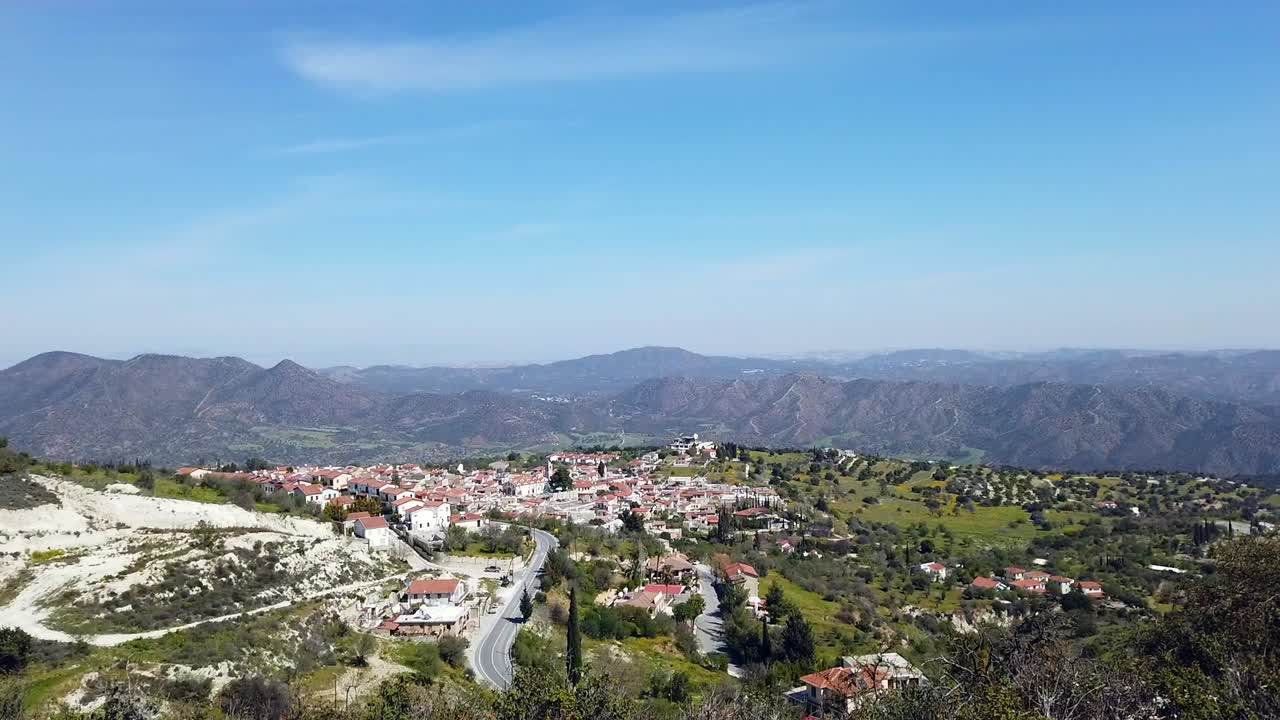 The camera pans from left to right capturing the beautiful village beside it, showcasing both in a stunning panoramic view. The peaceful hills and blue skies complete this breathtaking scene.