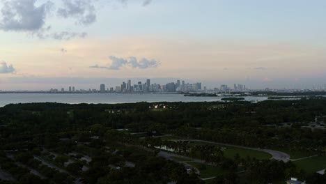 Dolly-Out-Toma-Aérea-Descendente-De-Un-Dron-De-La-Playa-Tropical-Rodeada-De-Palmeras-En-El-Parque-Crandon-En-Key-Biscayne-Con-El-Horizonte-De-Miami,-Florida-En-La-Distancia-En-Una-Soleada-Noche-De-Verano