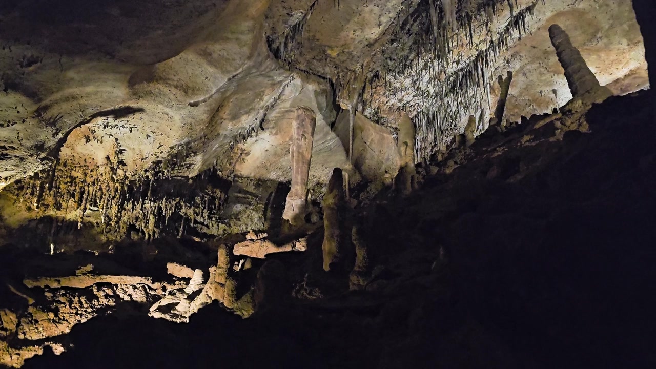 Wide view of a dark cave ceiling covered with stalactites and mineral deposits, with rocky ground, layered limestone walls, shadowy chambers, and warm guided tour lighting.