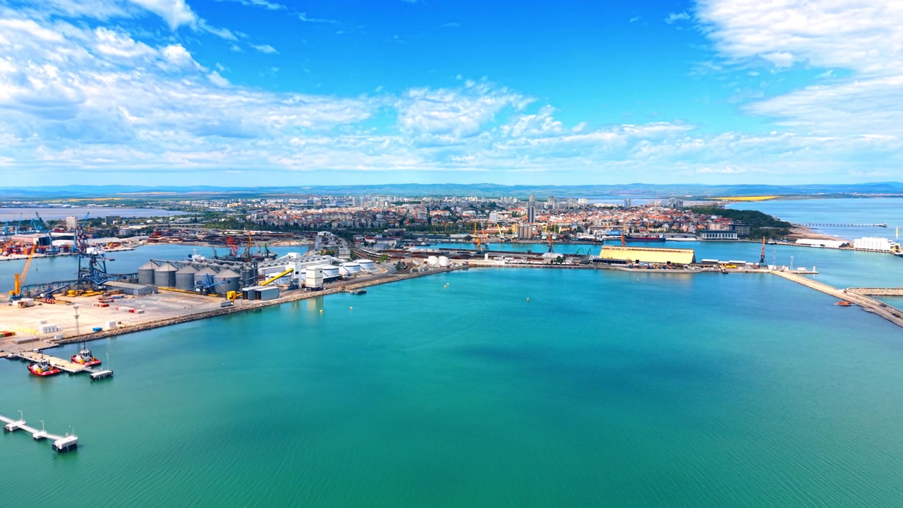 Wide aerial view of industrial harbor and turquoise sea. Aerial scenery of a large maritime port terminal with protective breakwater and city coastline.