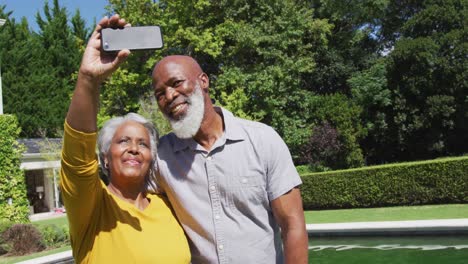 Happy-senior-african-american-couple-using-smartphone-taking-selfie-by-swimming-pool-in-sunny-garden