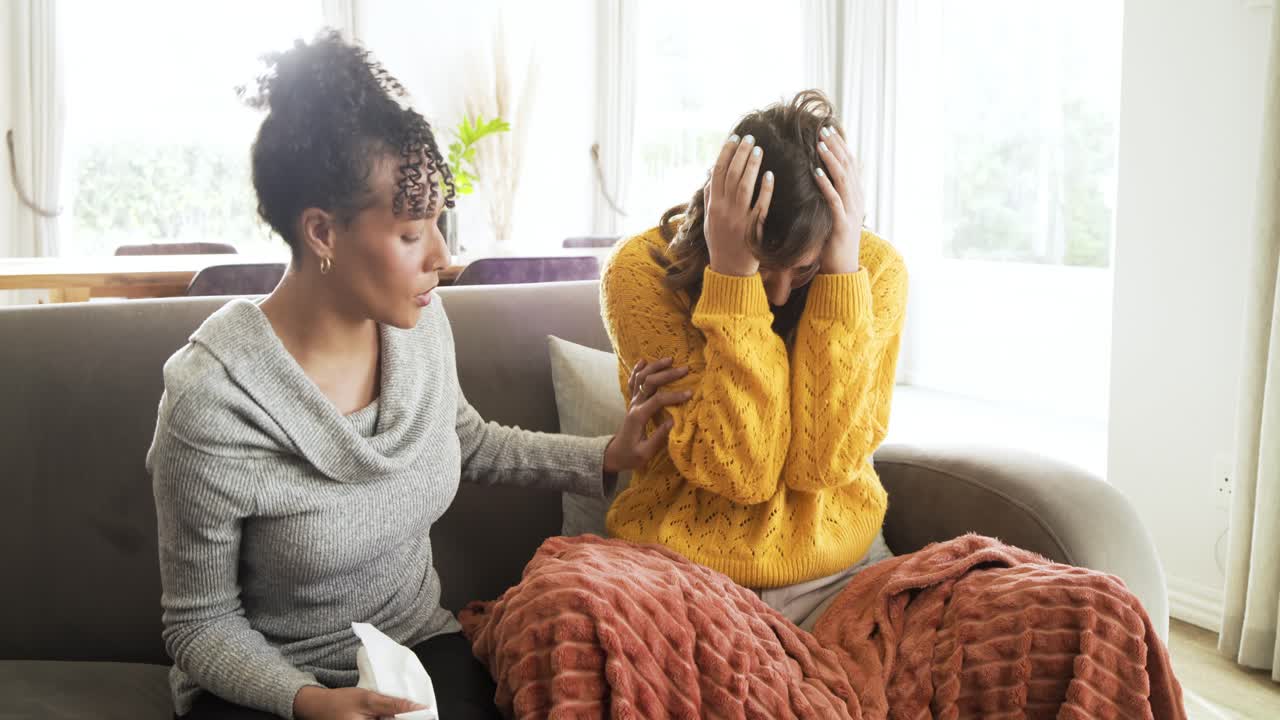 Diverse female friends reading smartphone message and offering tissues while sniffling on gray sofa