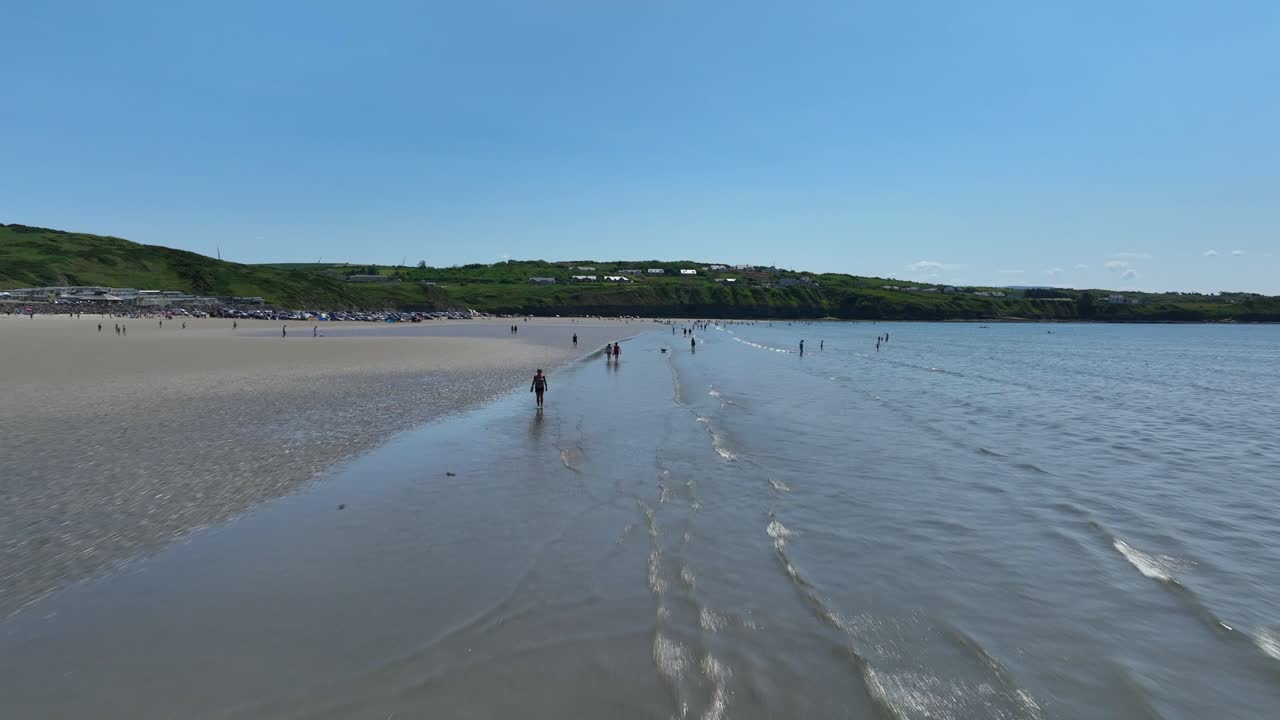 Rossnowlagh Beach, County Donegal, Ireland, June 2023. Drone pushes forward as waves roll in and people stroll along the strand with holiday cottages in the distance.