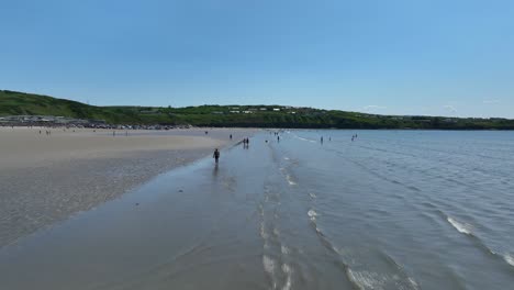 rossnowlagh beach, county donegal, ireland, june 2023. drone pushes forward as waves roll in and people stroll along the strand with holiday cottages in the distance.